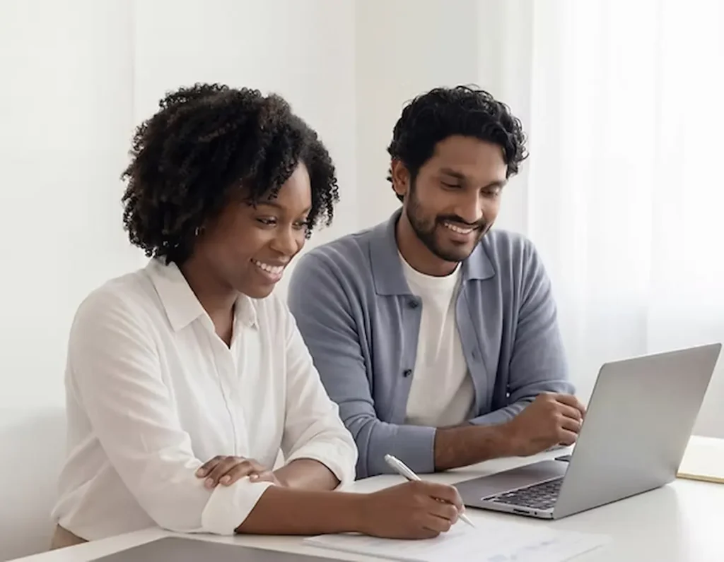 Two colleagues collaborating at a white desk. A woman wearing a white shirt smiles while writing notes on a piece of paper, sitting next to a smiling man in a blue shirt who is looking at a laptop screen.